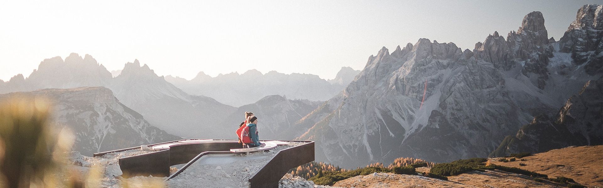 Due persone con zaini sono sedute su una piattaforma panoramica a forma di stella tra le montagne. Sullo sfondo si vedono cime rocciose e innevate delle Dolomiti, illuminate da una luce calda e bassa nel cielo.