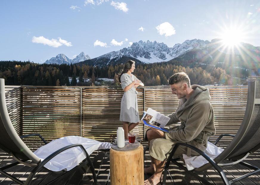 Ein Mann sitzt auf einem Schaukelstuhl und eine Frau steht am Balkon und genießt die Aussicht