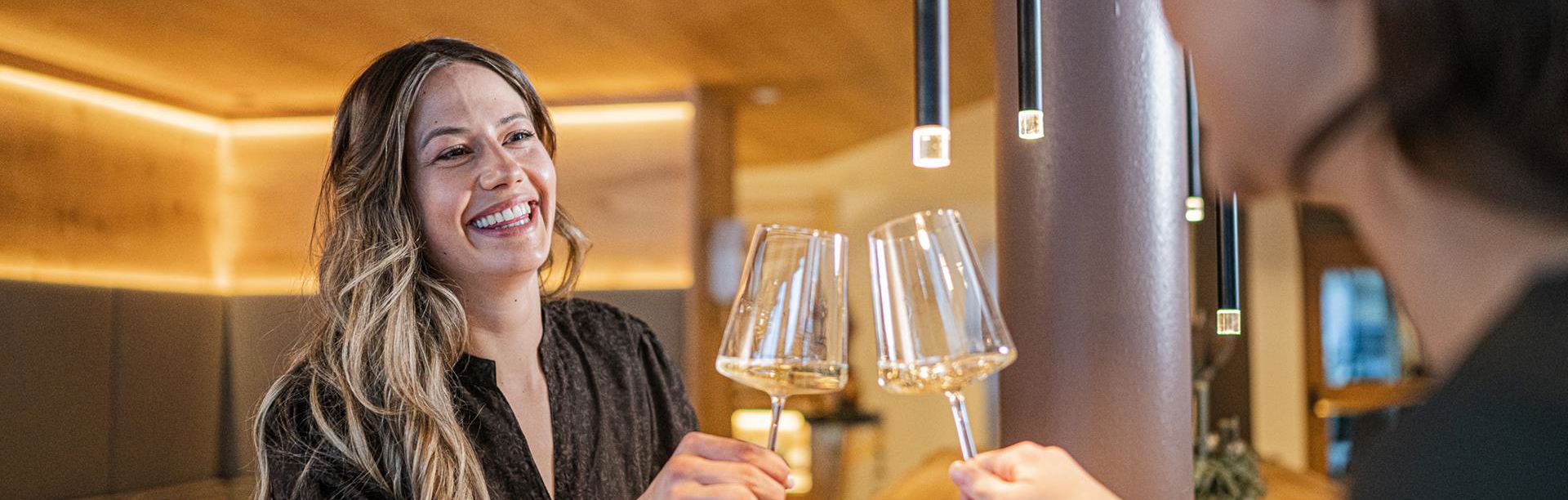 Due donne bevono un bicchiere di vino bianco; sul tavolo davanti a loro si trova un piatto di speck