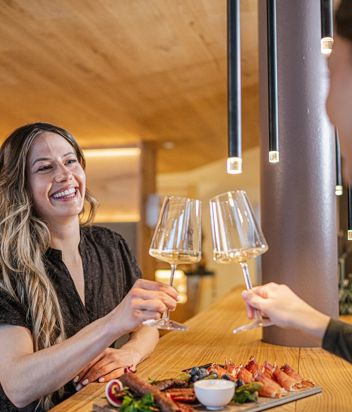 Zwei Frauen trinken ein Glas Weißwein, auf dem Tisch vor ihnen befindet sich eine Speckplatte