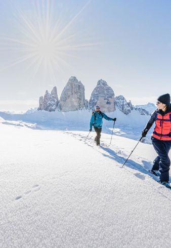 Zwei Personen sind beim Schneeschuhwandern unterwegs, im Hintergrund erheben sich die Drei Zinnen
