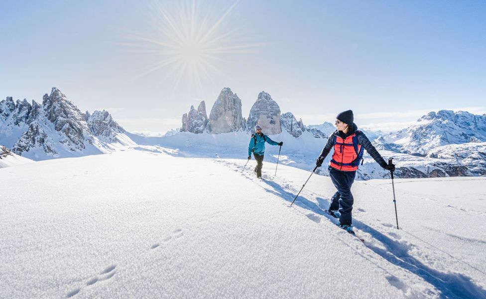Zwei Personen sind beim Schneeschuhwandern unterwegs, im Hintergrund erheben sich die Drei Zinnen