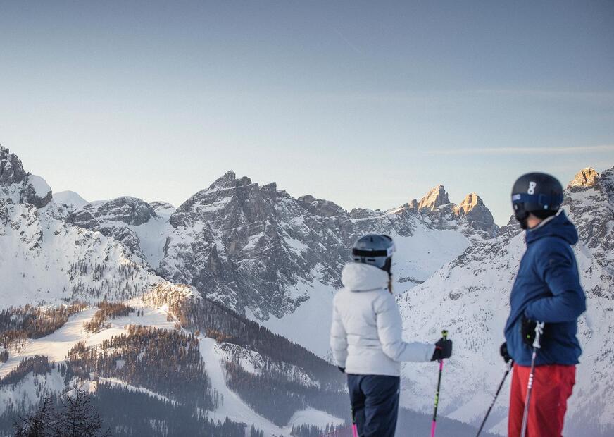 A man and a woman dressed in ski clothing look at the Dolomites in the Three Peaks ski area.