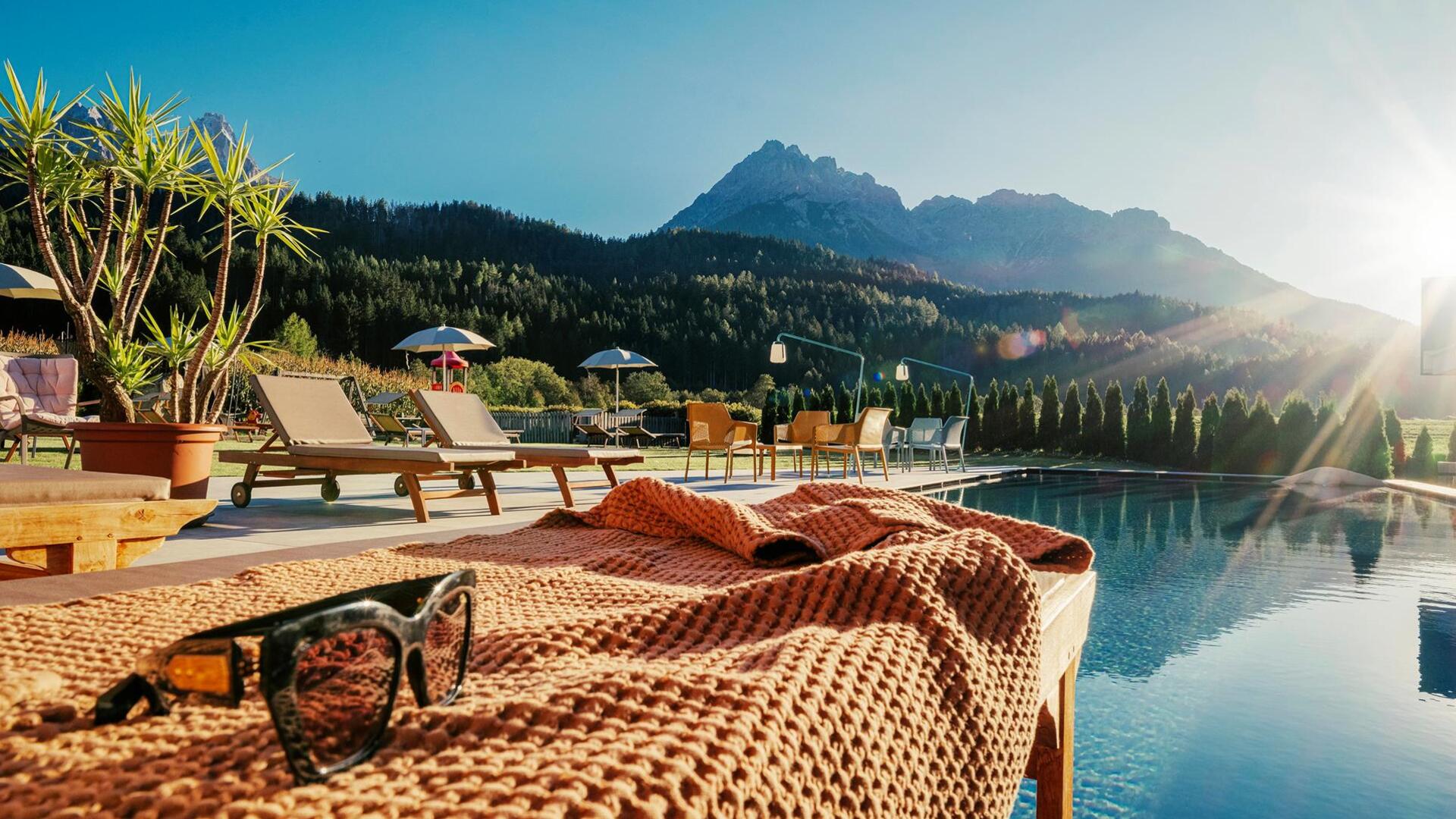 Eine Liege und Sonnenbrille am Poolrand mit anderen Liegen, Sonnenschirmen, Tischen und Stühlen und Haunold im Hintergrund