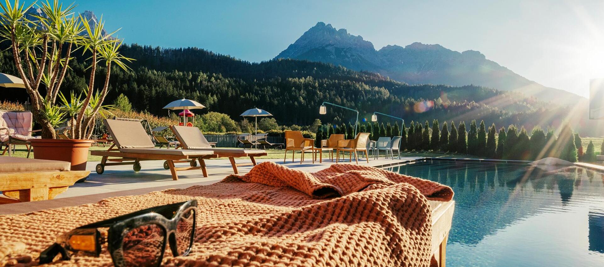 Eine Liege und Sonnenbrille am Poolrand mit anderen Liegen, Sonnenschirmen, Tischen und Stühlen und Haunold im Hintergrund