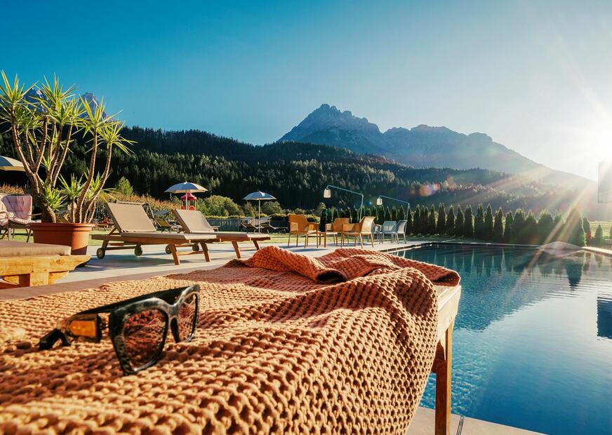 A sun lounger and sun glasses by the poolside with others sun loungers, sun umbrellas, small tables and chairs and the Haunold on the background