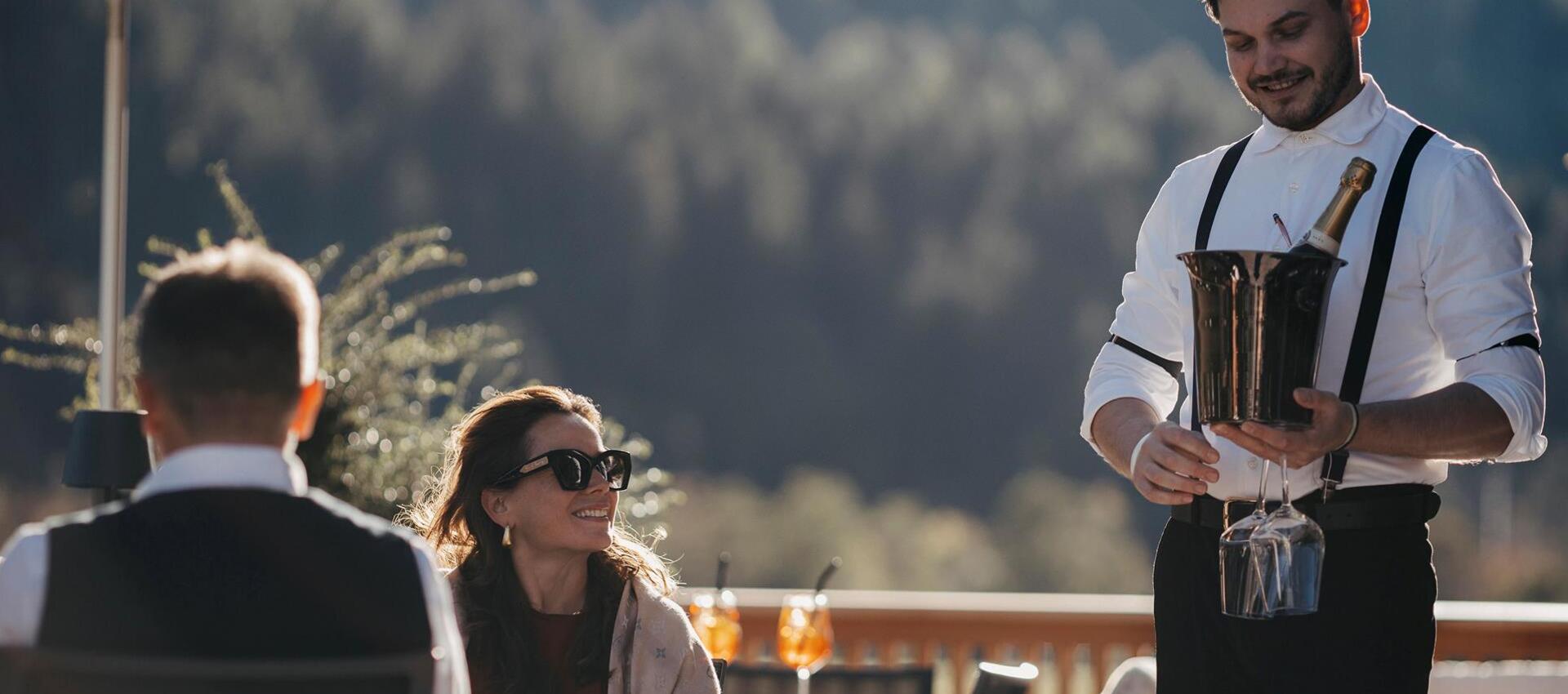 Un uomo porta due calici e una bottiglia di vino a un uomo e una donna seduti a un tavolo su una terrazza, sotto un ombrellone e con dietro un bosco
